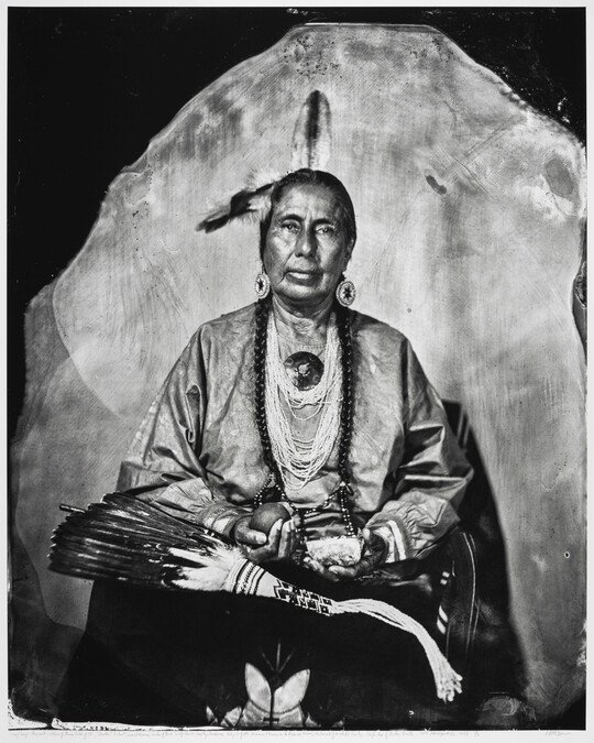 A black-and-white portrait photograph of a seated Indigenous person, feathers in their hair, strings of beads around their neck, holding several objects in their hands.