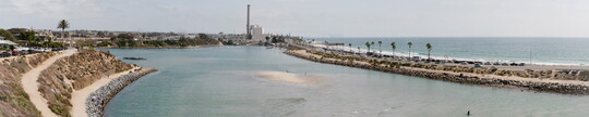 A panoramic color photograph of (from left to right) a rocky shoreline, a shallow bay, a narrow isthmus with traffic leading to a power plant, and a larger body of water.