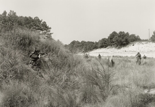 A black-and-white photograph of a person in military clothing crouching in tall grass and aiming a rifle at several other soldiers in a nearby field.