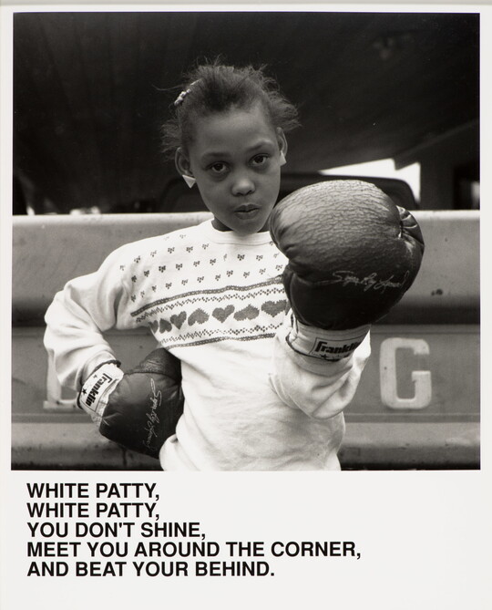A black-and-white photograph of a Black girl wearing boxing gloves, one hand on her hip the other aimed at the viewer, above text that reads, "White Patty, White Patty, You don't shine. Meet you around the corner and beat your behind."