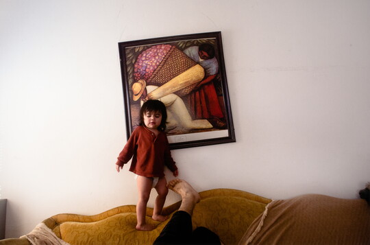 A color photograph of a toddler standing on the back of a couch looking down at a person whose leg is propped next to them, and a crooked artwork hanging above the couch.