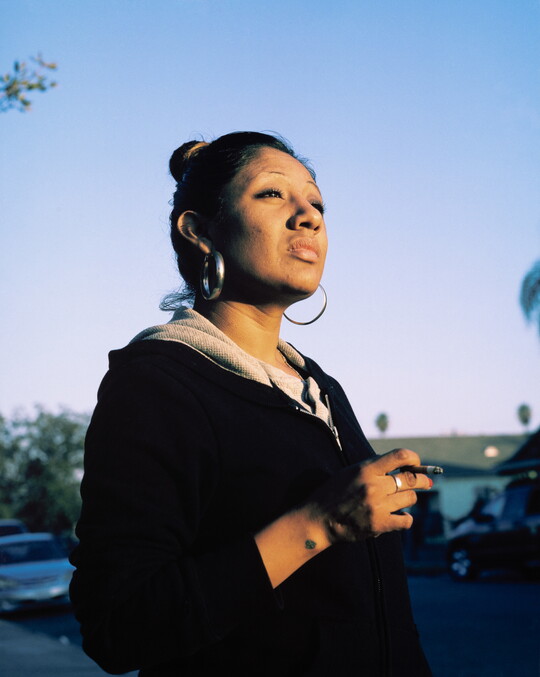 A color photograph of a medium-skinned woman, hair pulled on top of her head and large hoop earrings, standing outside holding a cigarette in her hand.