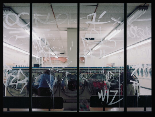 A color photograph of a laundromat at night through as seen through glass windows covered in white graffiti.