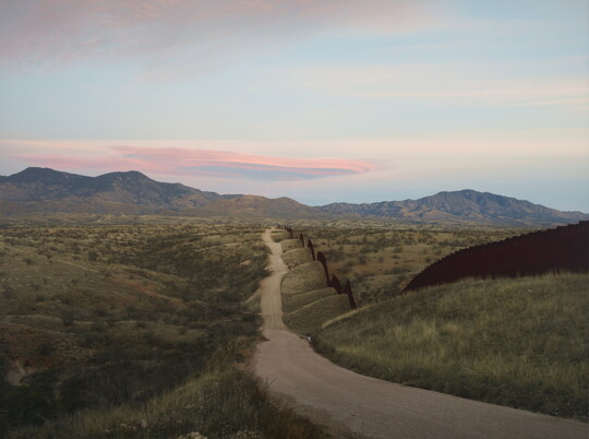 A color photograph of a dirt road that follows along a tall wall through rolling hills with mountains in the distance.
