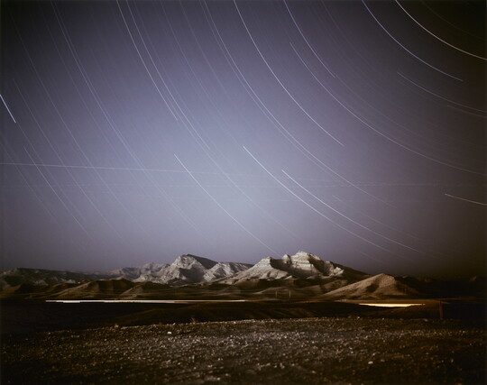 A color photograph of a dark desert landscape and mountains against a dark blue night sky with light streaks curving across the sky.