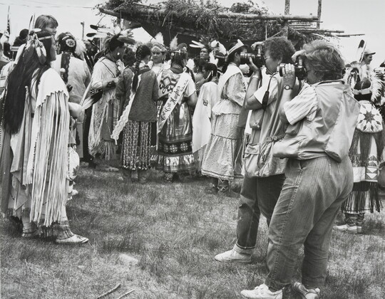 A black-and-white photograph of two women photographing a group of Indigenous people in cultural dress.