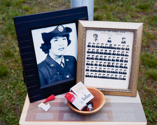 A color photograph of a table with two framed black-and-white photos: one of an Indigenous woman in military uniform, the other a class photo, and a wood bowl containing two packs of cigarettes and a lighter.
