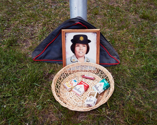A color photograph of a framed color photo of an Indigenous woman in military uniform behind a basket containing four packs of cigarettes and a lighter placed on grass.