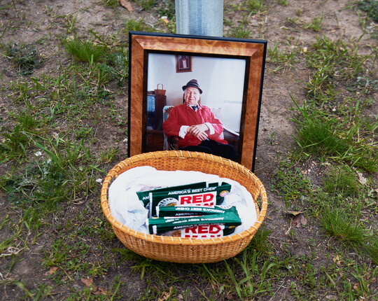 A color photograph of a framed color photo of a seated man in a red jacket behind a basket on grass filled with packets of chewing tobacco.