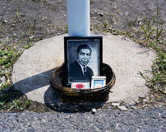 A color photograph of a woven basket on concrete containing a black-and-white framed photo of a man in a suit and two packs of cigarettes at the base of a pole.