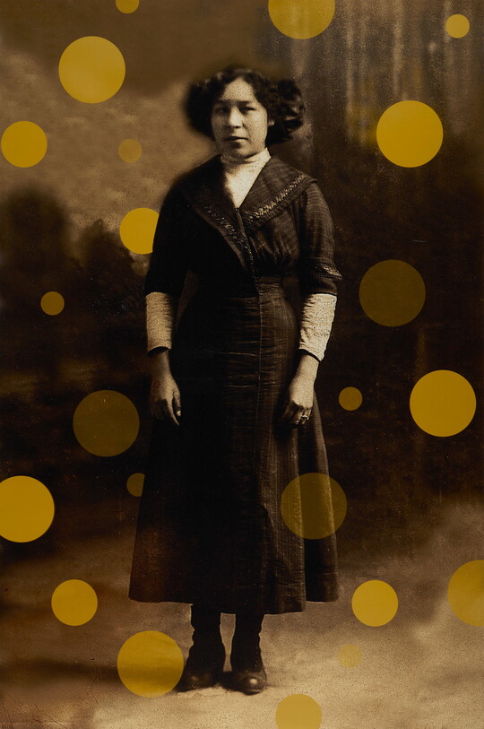 A sepia-toned portrait photograph of an Indigenous woman wearing an ankle-length A-line dress, hair cut short, with yellow dots floating around and behind her figure.