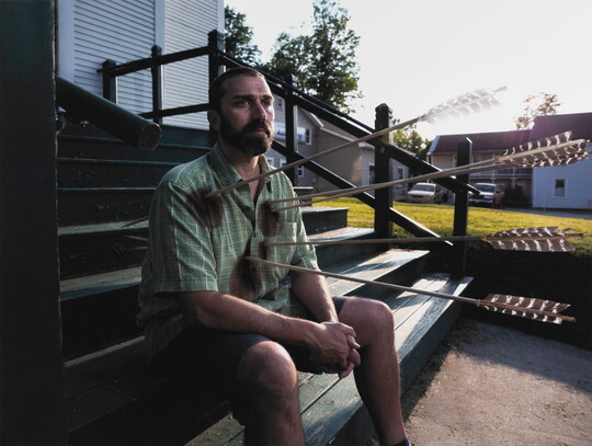 A color photograph of a White man sitting casually on wood steps with large arrows sticking out of his chest.