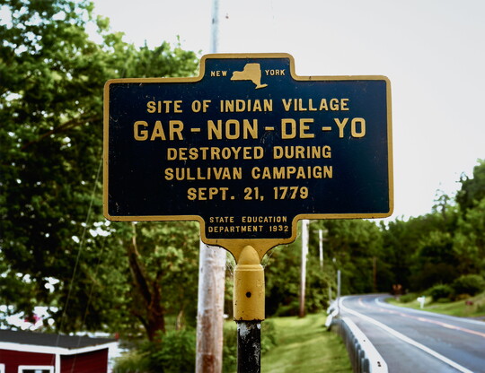 A color photograph of a New York historical marker that reads, "Site of Indian Village Gar-Non-De-Yo Destroyed during Sullivan Campaign Sept. 21, 1779," next to a road.