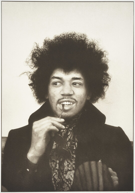 A black-and-white portrait photograph of a Black man with an Afro, looking to the side, holding a pair of sunglasses with the temple tip between his lips.