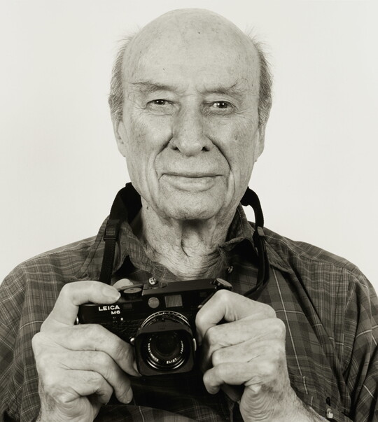 A black-and-white photograph of an older, White balding man from the chest up, looking at the viewer and holding a camera up under his chin.