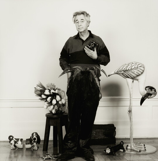 A black-and-white photograph of a White man standing with a camera in one hand, surrounded by plants and whimsical animal sculptures.