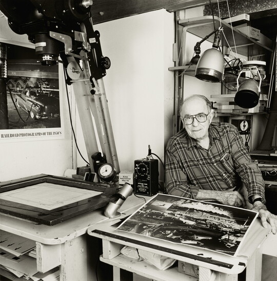 A black-and-white photograph of an older White man looking at the viewer from a photography studio work space.