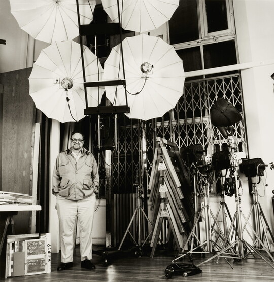 A black-and-white photograph of a balding White man standing with hands in pockets among large lighting umbrellas and other equipment.