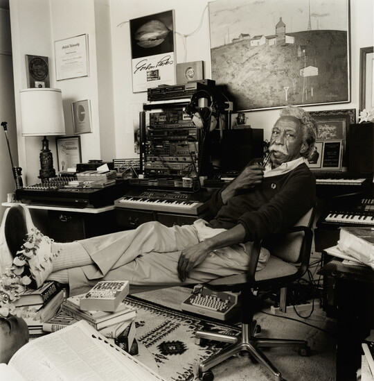 A black-and-white photograph of an older Black man with gray hair and a mustache holding a pipe to his lips, reclining in a chair in a cluttered office.