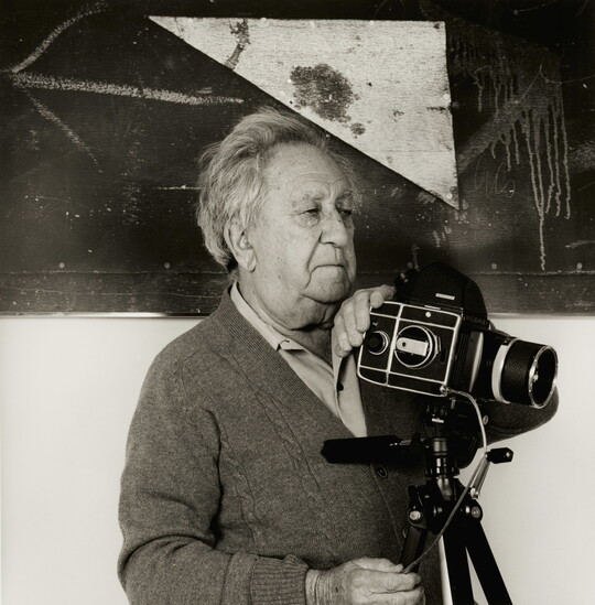 A black-and-white photograph of an older White man holding a camera on a tripod in front of a white and black wall.