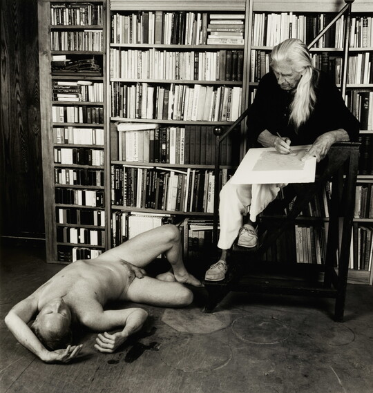 A black-and-white photograph of an older White man with long gray hair seated in front of a book shelf drawing a nude White man who is lying on the floor.