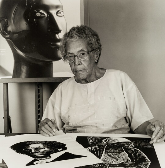 A black-and-white photograph of an older Black woman with two prints on a table in front of her and a large black sculpture of a head behind her.