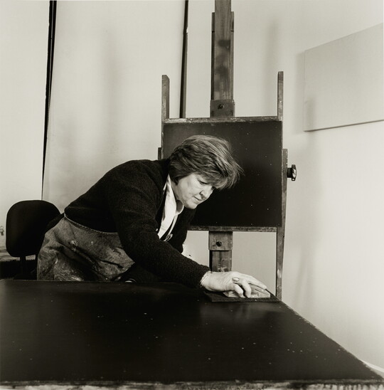 A black-and-white photograph of a White woman working on a large painting with an easel displaying a small painting behind her.