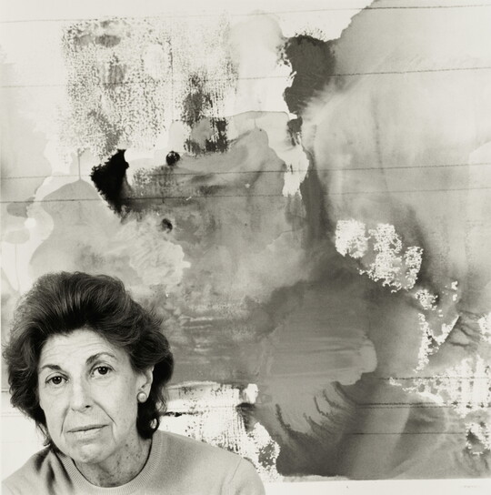 A black-and-white photograph of an older White woman, positioned in the lower left corner, in front of an abstract painting.