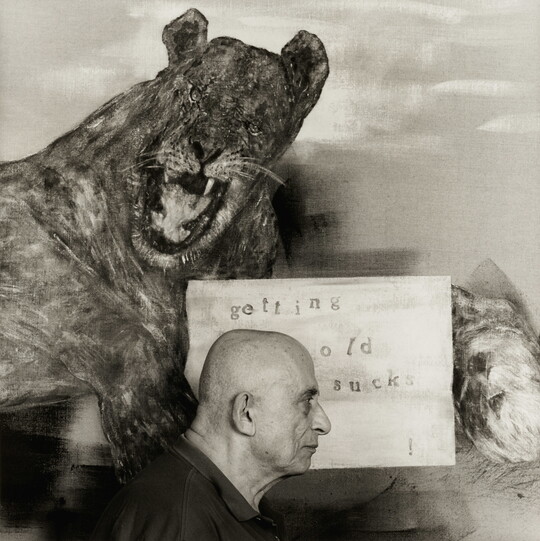 A black-and-white photograph of an older White man in profile standing in front of a painting of a lion holding a sign that says, "getting old sucks."
