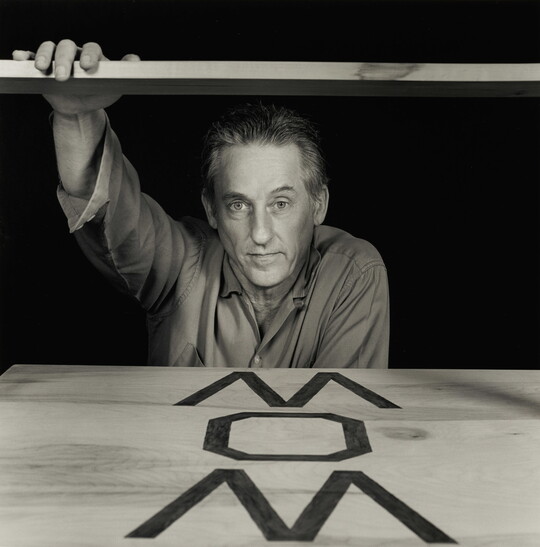 A black-and-white photograph of a White man hovering over a board with large letters written on it.
