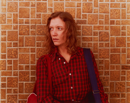 A color photograph of a White woman with reddish hair wearing a plaid, button-down shirt standing against a tile wall and looking to her right.