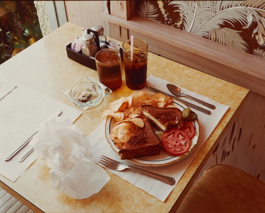 A color photograph of a dining table set with a full plate of food on a white paper placemat, full drinking cups, and an ashtray.