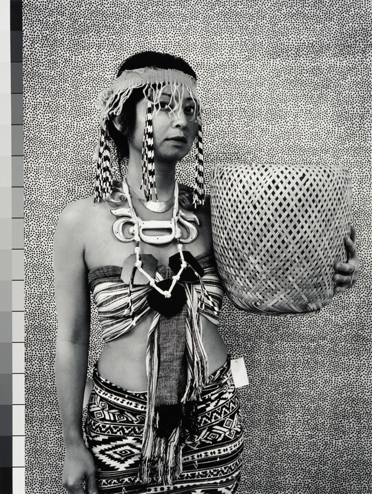 A black-and-white photograph of a medium-skinned woman wearing a headband with long beaded fringes, a halter top, patterned skirt, and holding a woven basket.