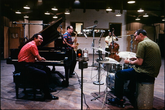 A color photograph of four men, three White and one Black, in a recording studio: one playing a piano, one a saxophone, one the drums, and one playing an upright bass.