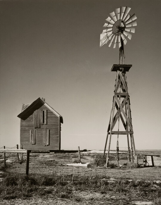 A black-and-white photograph of an abandoned boarded up farmhouse and a windmill.