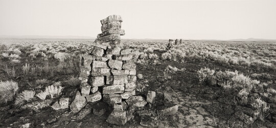A black-and-white photograph of several tower-like mounds of stones, receding into the distance, in an arid landscape dotted with sagebrush.