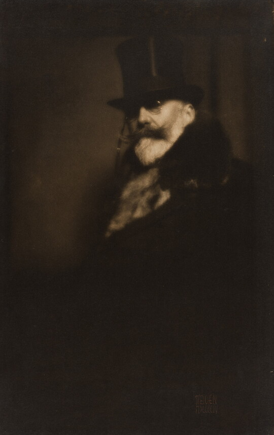A dark sepia-toned portrait photograph of a White man with a white beard and dark mustache, wearing a top hat, glasses, and a fur coat.