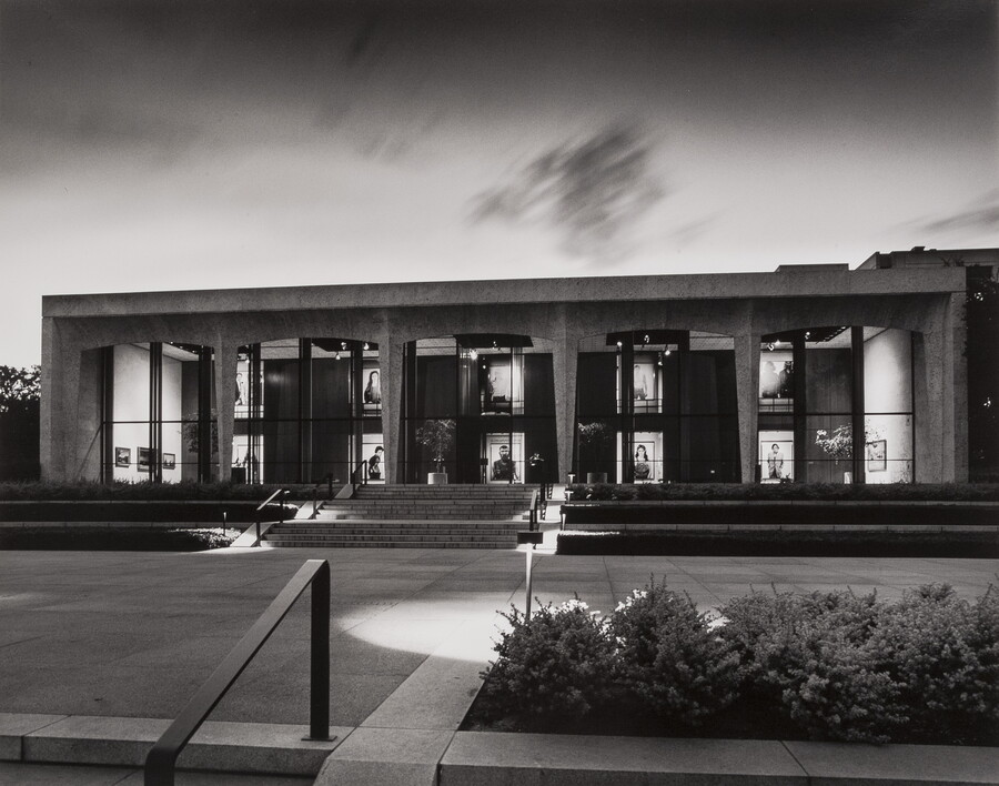A black-and-white photograph of the facade of the Carter at dusk; large-scale portrait photographs can be seen through the Carter's windows.