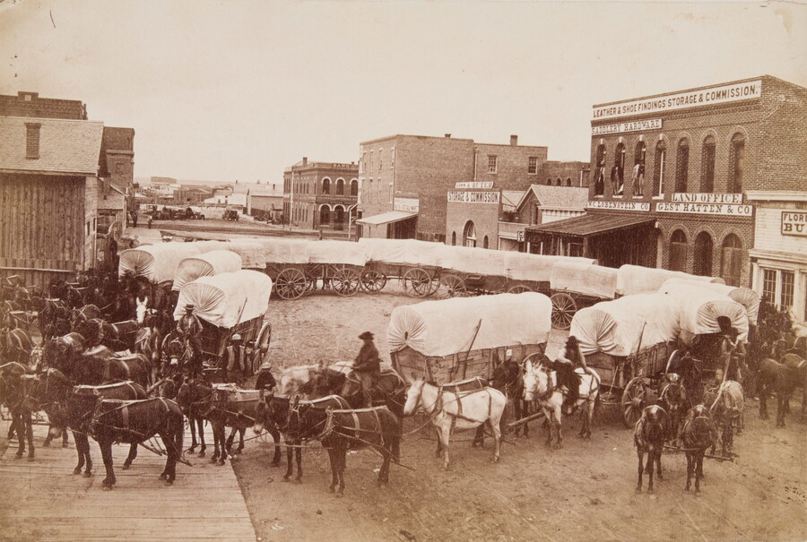 A sepia-toned photograph of covered wagons arranged in a large circle on a dirt street in a town.