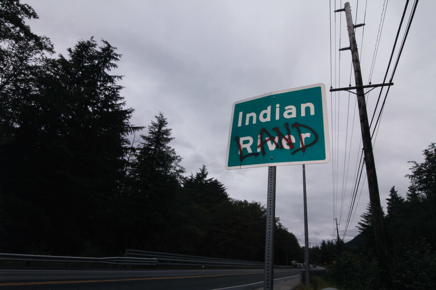 A color photo of a green road sign that says "Indian River" with the word "LAND" spray painted in red over "River."