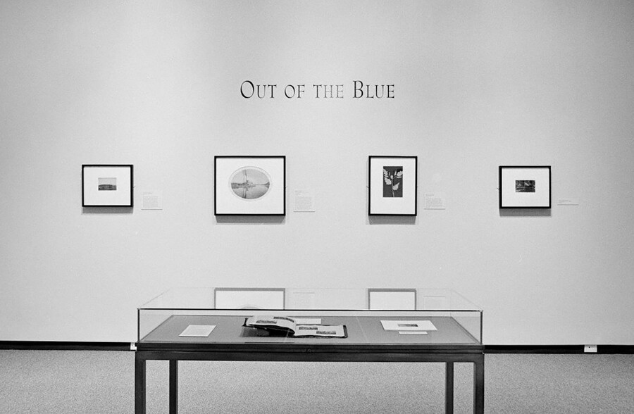 Black-and-white photo of a gallery; underneath the exhibition title, four framed artworks hang on a white wall; in the foreground is a display case containing an open book.