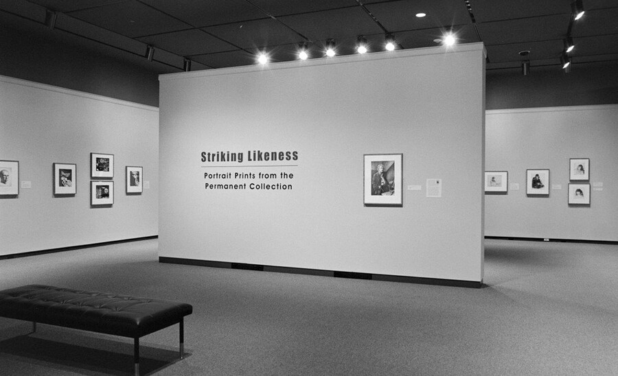 Black-and-white photo of gallery featuring the title wall with a framed portrait; in the background are more walls with framed artwork.