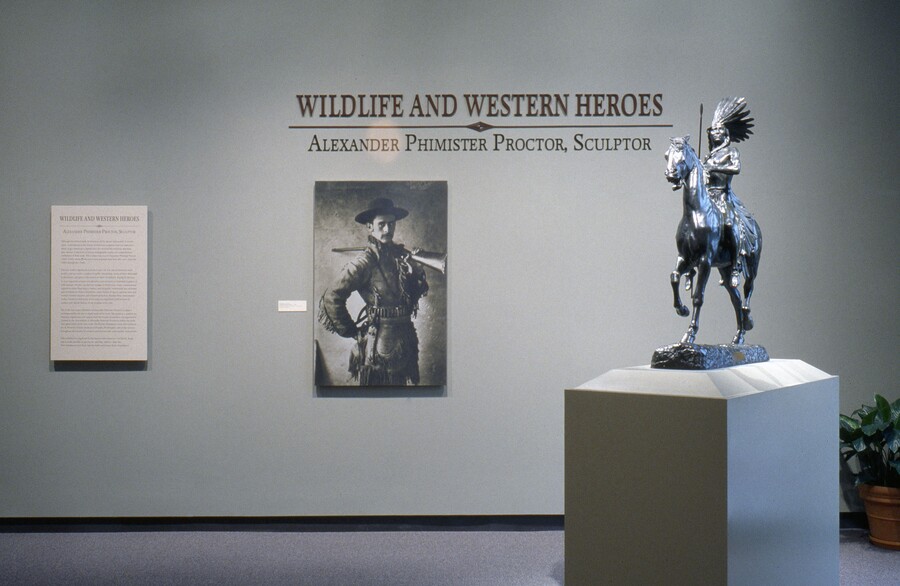 Under the title on a light gray wall is an enlarged photo of the artist dressed in cowboy garb; in the foreground on a pedestal is a sculpture of a Native American man riding a horse.