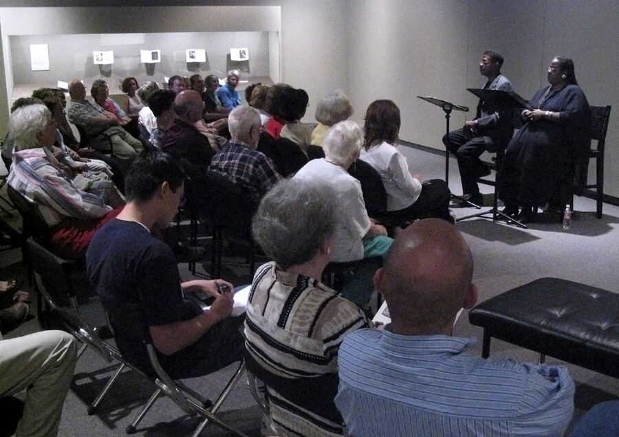 A crowd of people sit in a gallery in front of two performers who sit behind podiums.