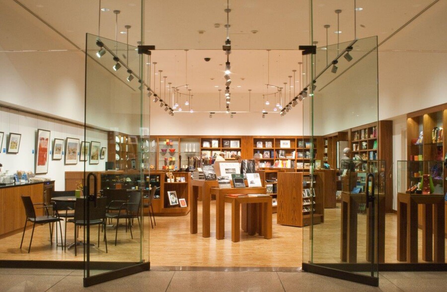 View of museum store from just outside the glass doors; wood shelves line the far and right walls and are filled with merchandise; the left wall features framed artwork; in the center are display tables and shelves featuring a variety of merchandise.