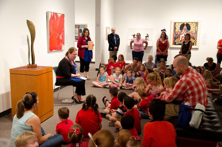 A woman sits in a gallery reading a book to children and adults gathered around her.