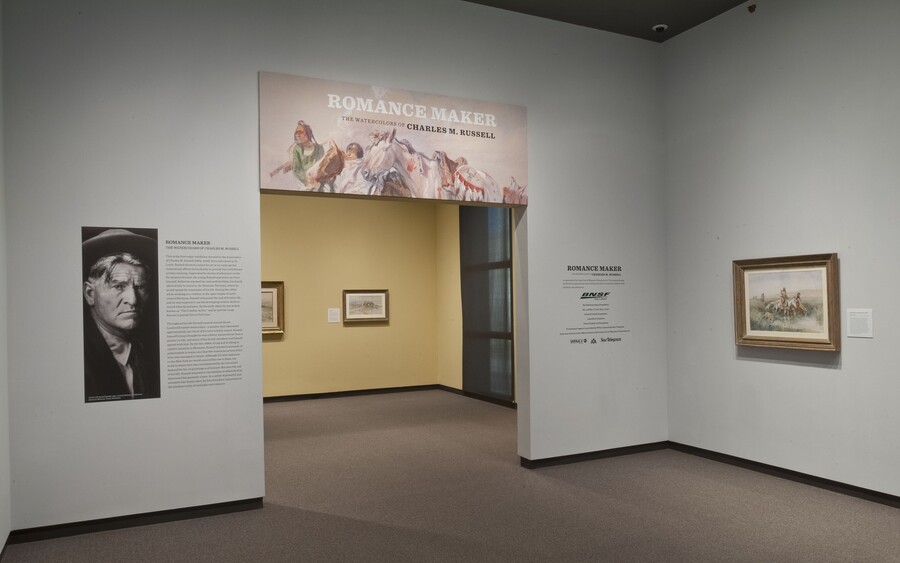 View of gallery title wall and entrance; to the left of the entrance is a large photo of the artist; over the entrance is the title graphic; on the wall to the right is a framed painting. More artwork can be seen through the entrance.