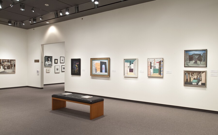 View of gallery showing framed paintings of different styles hanging on white walls. A black bench sits in front of some of the paintings.