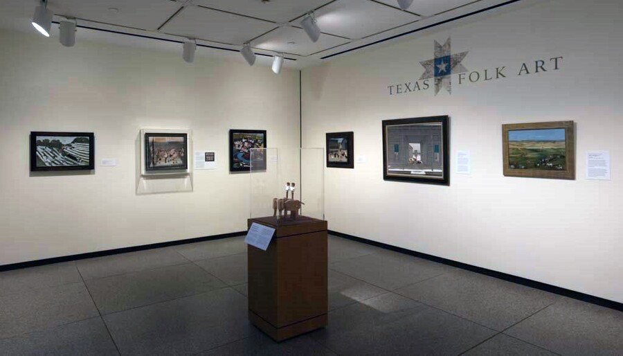 View of a gallery; both walls have three pieces of framed artwork hanging on them; above the artwork on the right wall is the exhibition title. In the foreground is a display case containing a sculpture.