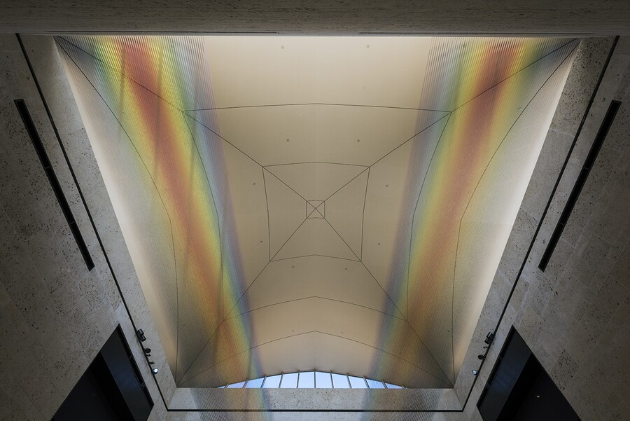 View of the ceiling of the museum&#039;s atrium showing the installation of two sections of colored thread stretched from wall to wall creating two rainbows.